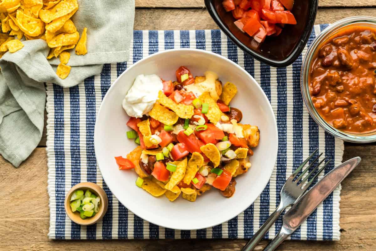 A bowl of Disney Totchos surrounded by toppings: chili, diced tomatoes, sliced green onions, and corn chips.
